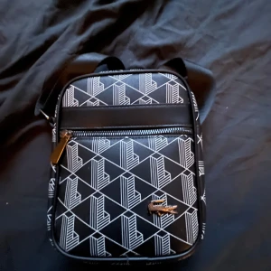 Svart axelväska från Lacoste med mönster - Säljer en svart axelväska från Lacoste med vitt geometriskt mönster och silverfärgad dragkedja. Väskan har en justerbar axelrem och klassisk Lacoste-logga framtill. Perfekt för att bära det viktigaste och ge din outfit en fet look