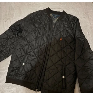  Ralph Lauren jacka - Snygg svart quiltad jacka från Ralph Lauren med dragkedja och ribbade muddar. Jackan har ett diskret broderat märke på bröstet och praktiska fickor med knappar. Perfekt för kyligare dagar.