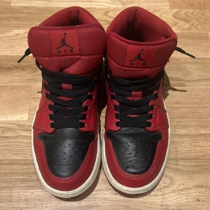 Röd-svarta Jordans 1s - Snygga Nike Air Jordans i rött och svart med klassisk design. Skorna har en hög modell med snörning och den ikoniska Nike-loggan på sidan. Perfekta för dig som vill ha en stilren och sportig look.