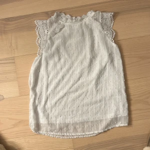 Vit ärmlös blus med spetsdetaljer - Säljer en söt vit blus med spetsdetaljer vid ärmarna. Blusen är ärmlös och har ett diskret prickigt mönster. Perfekt för en somrig look! 🌼