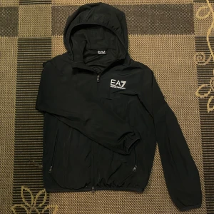 Svart Emporio Armani jacka Windbreaker - Säljer en stilren svart vindjacka från Emporio Armani med huva och dragkedja. Jackan har EA7-logga på bröstet och är perfekt för blåsiga dagar. Den är tillverkad i ett lätt syntetmaterial och har långa ärmar.Svart Emporio Armani jacka Windbreaker i riktigt bra skick som ändast är använd ett fåtal gånger! Vindjackan är äkta och har en scann på insidan! På insidan finns även en innerficka! frågor och funderingar, skriv direkt till mig! ⛓️‍💥 