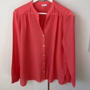 Blus i korall från Filippa K - Tunn och böljande korallfärgad blus från Filippa K. ”Crepe Shirt Blouse” V-ringad och med knäppning. Längd fram 65 cm, längd bak 70 cm. Vid i modellen. 
