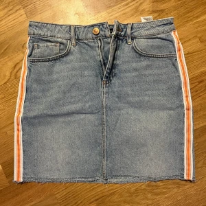 Jeanskjol från Bik Bok - Snygg jeanskjol från Bik Bok med orange och vita ränder längs sidorna. Kjolen har en klassisk femficksdesign och stängs med knapp och dragkedja framtill. Perfekt för en avslappnad stil.