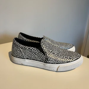 Svartvita slip-on sneakers från Zara - Snygga slip-on sneakers från Zara med ett svartvitt mönster, liknande ormskinn. Skorna har en vit sula. Storlek 38, normala i storleken. Mycket bra skick, endast lite smutsiga i sulan. 200kr eller bud.