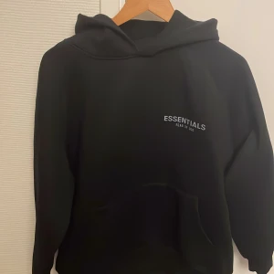 Svart hoodie från Essentials - Säljer en svart hoodie från Essentials, Fear of God. Den är superbekväm och perfekt för en skön stil. Hoodien har en klassisk design med en känguruficka framtill och Essentials-loggan tryckt på bröstet. Perfekt att ha hemma, i skolan eller ute.