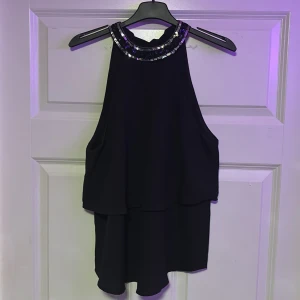 Svart ärmlös topp från ONLY - Snygg svart ärmlös topp från ONLY med glittrande detaljer runt halsen. Perfekt för en kväll ute eller en speciell tillställning. Toppen har en elegant och flytande design som ger en stilren look.