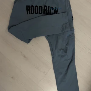 mjukisbyxor från Hoodrich - Säljer ett par blå mjukisbyxor från Hoodrich med svart text framtill. Byxorna har en avslappnad passform och är perfekta för en bekväm stil. De har elastisk midja och muddar vid bensluten.