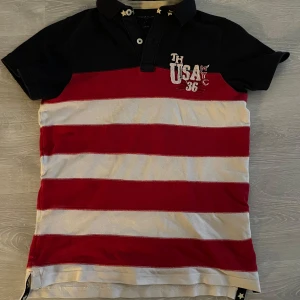 Pikétröja från Tommy Hilfiger med stars & stripes motiv - Säljer en snygg randig pikétröja från Tommy Hilfiger i rött, vitt och marinblått. Tröjan har korta ärmar och broderad text på både fram- och baksidan. Perfekt för en avslappnad stil med en touch av USA-inspiration.