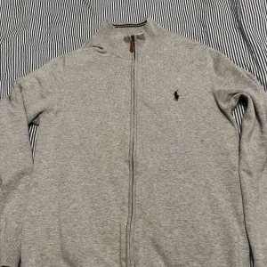 Grå kofta från Polo Ralph Lauren - Säljer en stilren grå kofta från Polo Ralph Lauren med dragkedja och broderad logga på bröstet. Koftan har en ribbad krage och är perfekt för en avslappnad men snygg look. Passar bra till både jeans och chinos. Den är i mycket bra skick 
