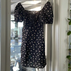 Blommig klänning i linneblandning - Säljer en söt blommig klänning i linneblandning med puffiga kortärmar och knytning framtill. Klänningen har en smockad rygg och ett romantiskt blommönster i rosa och grönt på en mörk bas. Perfekt för varma dagar!💕🌷