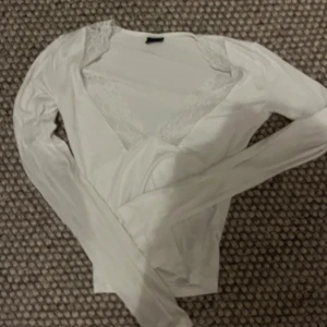 Vit långärmad blus med spetsdetaljer - Elegant vit blus med långa ärmar och v-ringad design. Blusen har vackra spetsdetaljer vid halsringningen som ger en romantisk touch. Perfekt för en stilren och sofistikerad look.