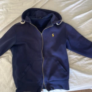 Mörkblå hoodie från Polo Ralph Lauren - Säljer en snygg mörkblå hoodie från Polo Ralph Lauren med dragkedja och det klassiska broderade logotypen i gult på bröstet. Tröjan har en justerbar huva med snören och fickor framtill. Perfekt för en avslappnad stil.
