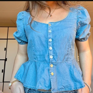 Blå blus med knappar - Charmig blå blus med korta puffärmar och knappar framtill. Blusen har en söt volangdetalj vid nederkanten som ger en feminin touch. Perfekt för en avslappnad och stilren look. Sitter som en s!💞