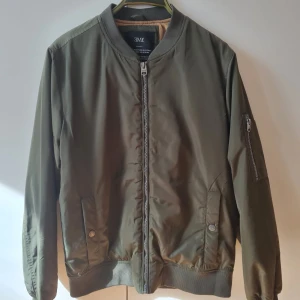 Khaki bomberjacka från Ellos - Snygg khaki färgad bomberjacka från Ellos med dragkedja framtill och ribbade muddar. Jackan har långa ärmar och praktiska fickor med knappar. Lättfodrad, finns innerfickor med dragkedja. Storlek S. Kan hämtas i Vallentuna.