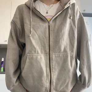 Grå hoodie med dragkedja - Säljer en bekväm grå hoodie med dragkedja och justerbar huva. Perfekt för en avslappnad stil. Den har fickor framtill.
