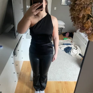 Svart oneshoulder topp - Snygg svart oneshoulder topp i ett elegant och glansigt material. Perfekt för en stilren look med en öppen rygg som ger en extra touch. Passar utmärkt till både byxor och kjol.