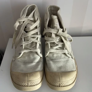 Off-white  sneakers från Palladium - Säljer ett par vita sneakers från Palladium med hög snörning och robust sula. Skorna är tillverkade i canvas och har en rund tå. Perfekta för en avslappnad stil.