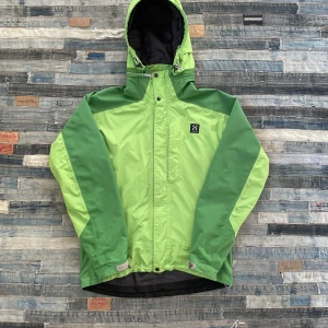 Grön vindjacka från Haglöfs - Säljer en grön vindjacka från Haglöfs med huva och dragkedja. Jackan har en tvåfärgad design med ljusare och mörkare gröna paneler. Perfekt för utomhusaktiviteter med justerbar huva och flera praktiska fickor.