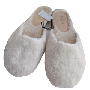 Vita fluffiga tofflor från Zign - Mysiga vita tofflor från Zign i ett fluffigt material som ger en varm och bekväm känsla. Perfekta för att hålla fötterna varma under kyliga dagar. De har en enkel slip-on design och en mjuk sula för extra komfort.
