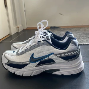 Vita och blå Nike sneakers - Snygga Nike sneakers i vitt med blå detaljer och en svart sula. Skorna har en klassisk snörning och en sportig design med Nike-loggan på sidan. Perfekta för en aktiv livsstil. Är helt nya men tyvärr för små  , pris går att diskuteras och pris går att sänkas vid snabbt köp 