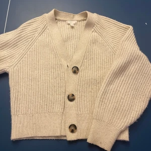 Krämvit ribbad kofta  - Säljer en stilren beige kofta med ribbad struktur och stora knappar framtill. Koftan har en v-ringad design och långa ärmar, perfekt för en avslappnad look. Jättemysig🥰