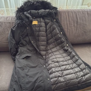 Svart parkajacka med pälskrage från [C]STUDIO - Säljer en stilren svart parkajacka från [C]STUDIO. Jackan har en mysig pälskrage och är quiltad på insidan för extra värme. Den har långa ärmar och stängs med både dragkedja och knappar. Perfekt för kyliga dagar! Passar för både M och L