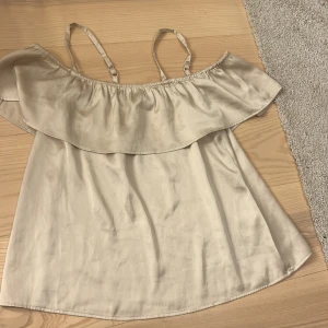 Beige offshoulder topp - Säljer en stilren beige offshoulder topp med volangdetalj. Toppen har smala axelband och är tillverkad i ett glansigt material som ger en lyxig känsla. Den har tock en liten blå fleck som inte går bort men det är inget man märker