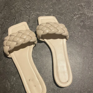 Beige sandaler med flätad rem - Snygga beige sandaler med en bred flätad rem över foten. De har en fyrkantig tå och en platt sula som ger en stilren look. Perfekta för varma sommardagar.