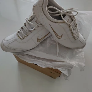 Vita sneakers från Nike (retro) - Snygga vita sneakers från Nike med gulddetaljer och snörning. Perfekta för en stilren look. Tillverkade i ÄKTA LÄDER av högsta kvalitet med en klassisk Nike-logga på sidan. En av dem första Nike modeller i mycket bra skick! Den verkliga storleken är 39!