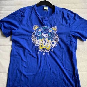 Blå t-shirt från Kenzo med tigertryck - Säljer en blå t-shirt från Kenzo med ett coolt tigertryck på framsidan och broderad logga på baksidan. T-shirten har en normal passform och är kortärmad, perfekt för en avslappnad stil.