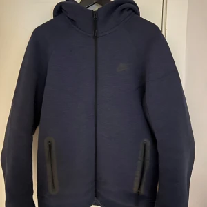 Nike tech - Använd 2 gånger, som ny. Inköpt för 1500kr. Säljer en mörkblå hoodie från Nike med dragkedja och huva. Jackan har långa ärmar och är tillverkad i polyester och bomull. Perfekt för en sportig look. Inga skador
