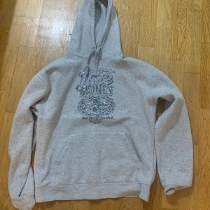 Grå hoodie med glittrig text - Säljer en grå hoodie med en glittrig textdesign på framsidan. Tröjan har en klassisk känguruficka och justerbar huva med dragsko. Perfekt för en avslappnad stil.