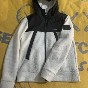  Sail racing jacka  - Säljer en snygg vit och svart fleecejacka från Sail racing . Jackan har en dragkedja framtill och en praktisk huva. Perfekt för kyliga dagar med sin mjuka och varma fleece. Den har långa ärmar och en stilren design.