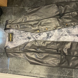 Svart skinnväst med knappar - Snygg svart skinnväst med knappar och en unik mönstrad insida. Perfekt för att ge din outfit en cool touch. Västen har en klassisk design med en stilren look.