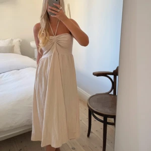 Beige axelbandslös klänning - Säljer en stilren beige axelbandslös klänning med knytband i halsen. Klänningen har en luftig passform och är perfekt för varma dagar. Den har en vacker rynkad detalj framtill som ger en extra touch.