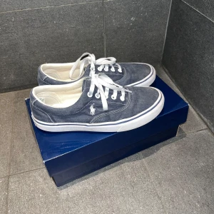 Blå tyg skor från Polo Ralph Lauren - Snygga blå sneakers från Polo Ralph Lauren med vit sula och snörning. Skorna har en klassisk design med märkets logga på sidan och insidan. Perfekta för en avslappnad stil. Öppet för bud. De är slitna på insidan men nästan ingenting på utsidan, nypris på dessa skor är runt 1000 kr.