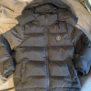 Svart pufferjacka från Trapstar - Säljer en svart pufferjacka från Trapstar med huva och dragkedja. Jackan har en broderad logga på bröstet och text på ryggen. Tillverkad i 100% polyester, perfekt för kalla dagar.