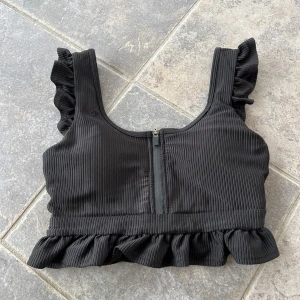 Aim’n Crop top small - Snygg svart ribbad topp med volangdetaljer vid axlar och nederkant. Toppen har en dragkedja bak och är perfekt för en trendig look.