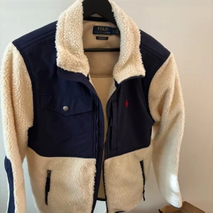 Vit teddyjacka från Polo Ralph Lauren - Säljer en snygg beige teddyjacka från Polo Ralph Lauren med marinblå detaljer. Jackan har en dragkedja framtill och två fickor med knappar. Perfekt för kyliga dagar med sin mysiga och varma design.