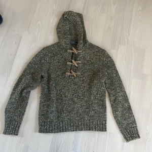 Gråmelerad stickad hoodie från Polo Ralph Lauren - Säljer en snygg gråmelerad stickad hoodie från Polo Ralph Lauren. Tröjan har en huva och unika träknappar framtill som ger en rustik känsla. Perfekt för kyliga dagar med sin mysiga och varma design. Tröjan är jävligt mjuk och skön! 