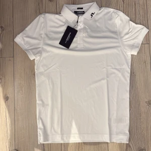  J.Lindeberg pike - Fräsch vit pikétröja från J.Lindeberg med svart logga på kragen och en snygg svart detalj över ryggen. Klassisk krage, korta ärmar och knäppning framtill. Tillverkad i mjuk polyester som andas, perfekt för en clean och sportig look. Regular fit, klassiskt Lent Sport material.