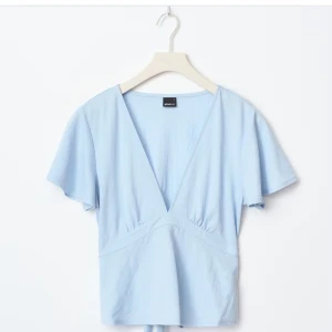 Blå tie top - Supersöt ljusblå omlottopp med v-ringning och knyt i midjan. Toppen har korta, lite fladdriga ärmar och markerad midja som ger en snygg siluett. Perfekt att styla med jeans eller kjol för en fräsch look.