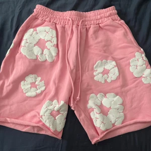 Rosa shorts från Denim Tears med blommor - Säljer ett par rosa shorts från Denim Tears med vita, upphöjda blommönster över hela plagget. De har elastisk midja med snörning och är tillverkade i mjuk bomull. Perfekta för dig som gillar statement-plagg och vill sticka ut med cool design.