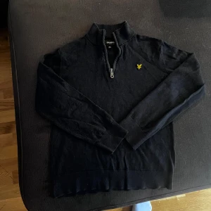 Svart half zip tröja från Lyle & Scott - Svart långärmad tröja från Lyle & Scott med half zip och klassisk gul logga på bröstet. Tröjan är i mjukt material och har en stilren, enkel design som passar perfekt till jeans eller chinos.