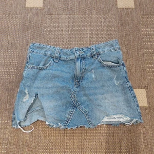 Blå jeanskjol från Gina Tricot - Snygg blå jeanskjol från Gina Tricot med slitna detaljer och rå kant. Kjolen har klassiska fickor fram och bak, bälteshällor och knappstängning. Perfekt för dig som gillar en cool och avslappnad stil.