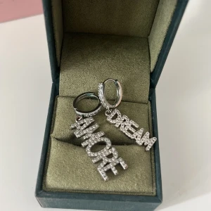 Glittriga örhängen med text - Säljer ett par coola silverfärgade örhängen med hängande texten 'AMORE' och 'DREAM' i stora bokstäver. Örhängena är täckta av glittrande stenar som ger extra bling och attityd. Perfekt för dig som vill sticka ut och sprida good vibes.
