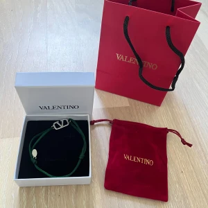 Grönt Valentino armband med silverdetalj - Snyggt armband från Valentino med grön textilrem och en stilren silverfärgad logodetalj. Kommer med original ask och röd sammetspåse med guldfärgad logga. Perfekt accessoar för dig som gillar exklusiva märken och stilren design.