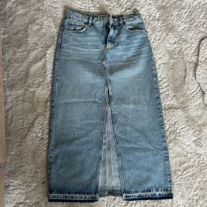 Lång jeanskjol med slits - Säljer en snygg lång jeanskjol i klassisk blå denim. Kjolen har en rak passform och en tydlig slits framtill som ger en cool vibe. Fickor både fram och bak samt hällor för bälte. Perfekt för dig som gillar streetstyle och vill ha något unikt i garderoben.