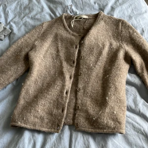 Beige kofta i ull från Gina Tricot - Mysig beige kofta från Gina Tricot i storlek XS. Koftan är tillverkad i ull och har en klassisk rund halsringning samt knappar framtill. Perfekt för lager-på-lager under kyliga dagar och har en enkel, tidlös look.