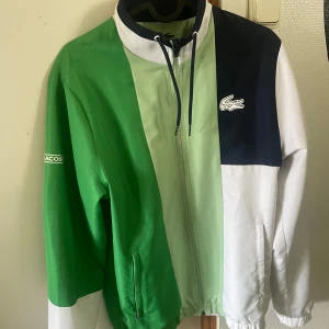 Lacoste tracksuit - Säljer ett par fräscha mjukisbyxor från Lacoste i vitt med gröna och mörkblå detaljer. Byxorna har resår i midjan och vid bensluten, samt en liten Lacoste-logga på låret. Perfekta för chill eller sportig stil.